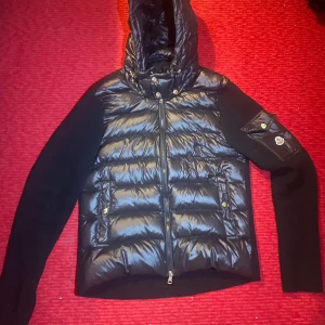 Svart Moncler cardigan - Snygg svart pufferjacka från Moncler med glansig front, stickade ärmar och huva. Jackan har dragkedja, fickor med tryckknappar och Moncler-logga på ärmen. Perfekt för dig som vill ha en stilren och varm jacka med streetkänsla.Size M amen lite M