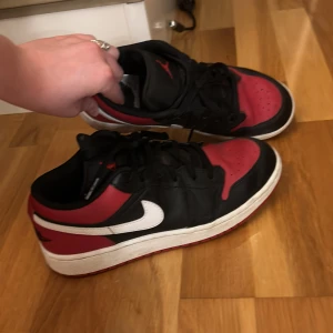 Nike Air Jordan 1 Low svart/röd - Säljer ett par Nike Air Jordan 1 Low med klassisk svart och röd färgkombination, vit swoosh och röd yttersula. Skorna har snörning och är tillverkade i skinn med perforerad tå för extra ventilation. Perfekta för dig som gillar streetstyle och sneakers med ikonisk design.