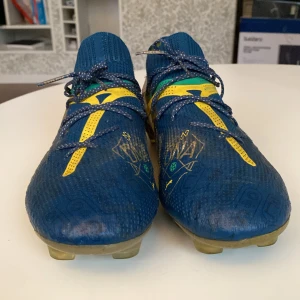 Blå-guld Puma fotbollsskor - Säljer ett par riktigt snygga Puma fotbollsskor i blått med guldiga och gula detaljer samt vita Puma-loggor. Det är Puma ultimate edition. Skorna har snörning, platt sula med dobbar och en stickad ovandel för skön passform. De har dock ritat med bläck på de vita puma loggorna, men det försvinner helt och hållet efter några fotbollsmatcher förhoppningsvis. De kostade 2899 kr när jag köpte de.
