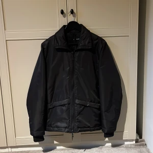 Svart bomberjacka från J.Lindeberg - Säljer en svart bomberjacka från J.Lindeberg i storlek M. Jackan har en clean look med dragkedja framtill, två snedställda fickor och hög krage. Materialet är glansigt och känns lätt men ändå värmande, perfekt för kyliga dagar.