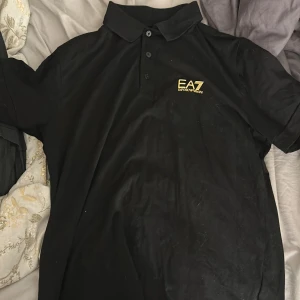 Svart EA7 Emporio Armani piké M - Svart pikétröja från EA7 Emporio Armani med klassisk krage och tre knappar framtill. Gult EA7-tryck på bröstet och gul detalj på ryggen. Tillverkad i mjuk bomull, kortärmad och med en clean, stilren look.