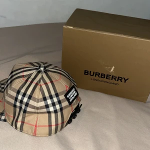 Burberry rutig keps i beige och svart - Snygg keps från Burberry med klassiskt rutigt mönster i beige, svart, vitt och röda linjer. Kepsen har en böjd skärm och justerbar knäppning bak. På baksidan sitter en svart patch med texten 'Burberry London England'. Perfekt accessoar för en trendig look.