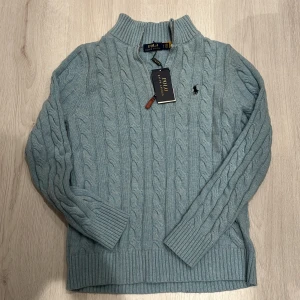 Ljusblå kabelstickad tröja Polo Ralph Lauren - Kabelstickad ljusblå tröja från Polo Ralph Lauren med halv dragkedja och ribbade muddar. Klassisk broderad logga på bröstet och hög krage. Perfekt för dig som gillar stilren och tidlös design.