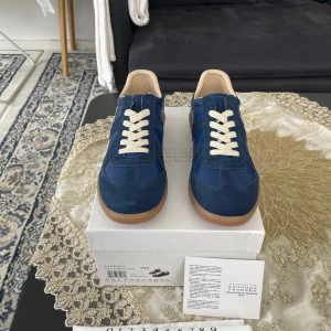 Maison Margiela blå sneakers - Snygga sneakers från Maison Margiela i marinblå mocka och skinn med vita snören och klassisk gummisula. Ikonisk design med diskreta detaljer och beige innersida. Perfekta för dig som vill ha en stilren och trendig look.