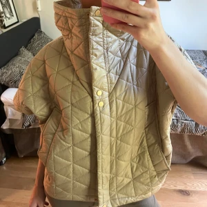 Beige quiltad pufferjacka - Säljer en beige quiltad pufferjacka med hög krage och tryckknappar framtill. Jackan har korta ärmar och oversized passform, perfekt för lager-på-lager. Materialet är syntet och jackan har ett snyggt rutmönster över hela ytan. Fickor på sidorna för extra funktion.