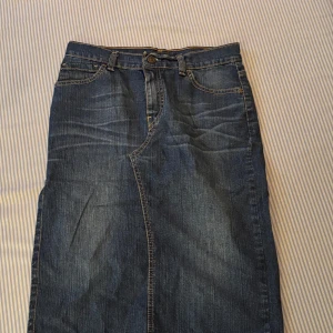 Lång jeanskjol från Levi's med slits - Säljer en klassisk lång jeanskjol från Levi's i mörkblå denim. Kjolen har fem fickor, bälteshällor och en snygg slits bak för extra rörelsefrihet. Perfekt till sneakers eller boots och enkel att styla med din favorit-topp.