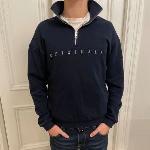 Marinblå half zip tröja Origianls - Snygg marinblå tröja från jack&jones med half zip och hög krage. Broderad text framtill och ribbade muddar. Perfekt för dig som gillar en clean och stilren look. Tillverkad i mjuk bomull för extra komfort.