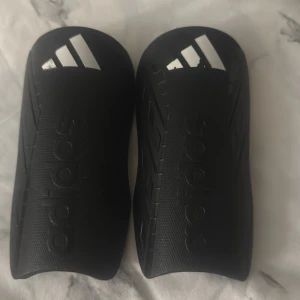 Svarta benskydd från Adidas - Säljer ett par svarta benskydd från Adidas med den klassiska loggan i vitt. De är tillverkade i tålig plast och har en snygg, sportig design med diskreta detaljer längs sidorna. Perfekta för fotbollsträning eller match.