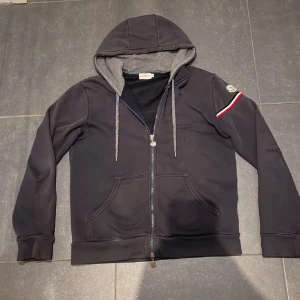 Mörkblå hoodie från Moncler zip up - Tja säljer nu en mörkblå hoodie från Moncler med grått foder i huvan, hel dragkedja och klassisk logga på ärmen. Snygg detalj med röd, vit och blå rand på ena ärmen. Känguruficka framtill och justerbara snören i huvan. Perfekt för en chill och stilren look. 