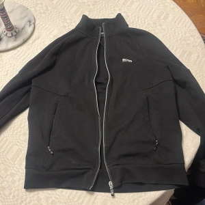 Svart ziptröja från Hugo Boss - Small men passar medium också. Svart ziptröja från Hugo Boss med vit logga på bröstet och coolt grafiskt mönster längs ärmarna. Tröjan har hög krage, två fickor med dragkedja och hel dragkedja framtill. Perfekt för dig som gillar stilren streetwear med snygga detaljer. 