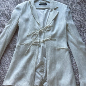 Vit kavaj med snörning från Gina Tricot - Snygg vit kavaj från Gina Tricot i storlek S. Kavajen har en krispig struktur, långa ärmar och två fickor framtill. Den stängs med två dekorativa snörningar och har en stilren, rak passform. Perfekt för dig som gillar minimalistisk och trendig stil.