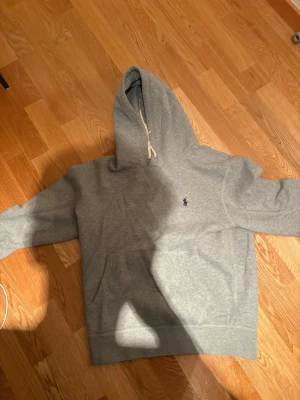 Grå hoodie från Polo Ralph Lauren - Snygg grå hoodie från Polo Ralph Lauren med klassisk huva och dragsko. Mjuk bomullsblandning och känguruficka framtill. Liten broderad logga i mörkblått på bröstet. Perfekt för en chill och stilren look. Inga defekter och är i väldigt bra skick 