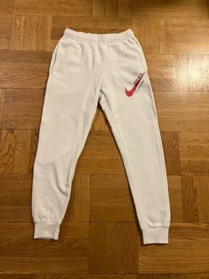 Vita Nike mjukisbyxor med röd logga - Säljer ett par vita mjukisbyxor från Nike med röd Swoosh-logga och text på vänster lår. Byxorna har resår i midjan och muddar vid bensluten för en skön och avslappnad passform. Perfekta för chill dagar eller träning.