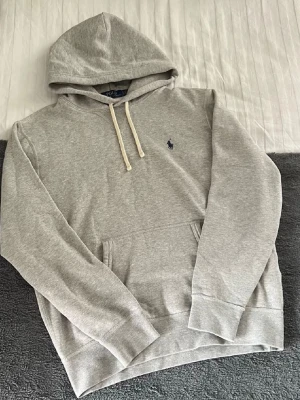 Grå hoodie från Polo Ralph Lauren - Snygg grå hoodie från Polo Ralph Lauren med klassisk huva och vita dragsnören. Tröjan har en känguruficka framtill och den ikoniska lilla loggan broderad på bröstet. Tillverkad i mjukt bomullsmaterial som är perfekt för chill dagar.