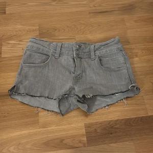 Grå jeansshorts med rå kant - Säljer ett par grå jeansshorts med rå, fransig kant och klassisk femficksdesign. Shortsen har knappgylf och bälteshällor, perfekta för en avslappnad stil. Passar dig som gillar en edgy look.