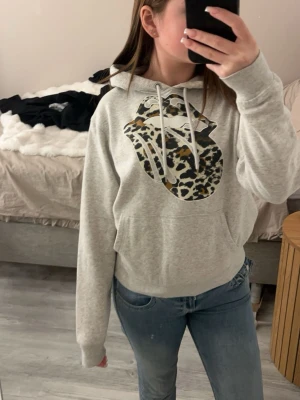 Grå hoodie med leopardmönstrad tryck - Snygg grå hoodie med huva och dragsnören. På bröstet finns ett stort tryck av Rolling Stones-loggan med leopardmönster. Hoodien har en klassisk känguruficka framtill och är tillverkad i mjukt material, perfekt för en avslappnad stil.