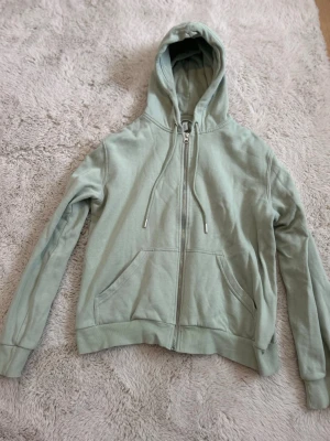 Ljusgrön hoodie med dragkedja - En stilren ljusgrön hoodie med dragkedja framtill och snörning i huvan. Hoodien har två stora fickor fram och ribbade muddar vid ärmar och nederkant. Jag har använt det mycket men säljer den eftersom att den börjar bli lite liten.