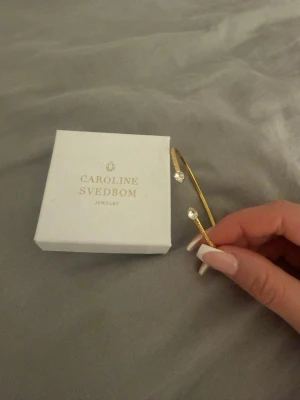 Guldarmband Caroline Svedbom - Snyggt och stilrent armband från Caroline Svedbom i gul med två gnistrande kristaller i ändarna. Öppen design som gör det lätt att ta på och av. Perfekt accessoar för att lyfta din outfit med lite extra bling. Använt 1 gång!