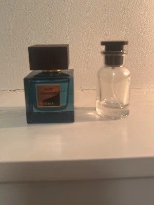 Blå parfymflaska med svart lock rituals  - Snygg parfymflaska i blått glas med fyrkantig form och svart lock, samt en genomskinlig mindre flaska bredvid. Flaskan har en guldfärgad etikett framtill och ger ett lyxigt intryck. Perfekt för dig som gillar stilrena och moderna doftflaskor.