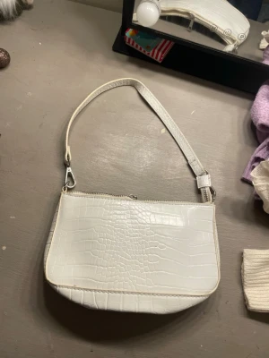 Vit croc handväska med dragkedja - Snygg vit handväska i croc-mönstrat syntetmaterial. Väskan har ett kort axelband och dragkedja upptill samt en praktisk ytterficka med dragkedja på baksidan. Perfekt accessoar för att lyfta din outfit.