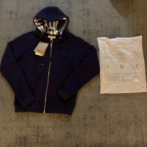 Marinblå Burberry hoodie med dragkedja - Snygg marinblå hoodie från Burberry med klassiskt rutigt foder i huvan. Tröjan har dragkedja framtill, snörning i huvan och ribbade muddar. Perfekt för dig som vill ha en stilren och exklusiv look med ikoniska Burberry-detaljer.                                                                                                                 Obs!  Originaltaggar medföljer ej