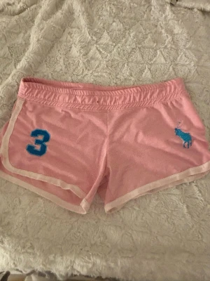 Rosa shorts från Polo Ralph Lauren - Snygga rosa shorts från Polo Ralph Lauren med vit kant, blå broderad logga och siffran 3 på ena benet. Perfekta för en sportig och avslappnad stil. Mjukt material och resår i midjan för extra komfort., snöret är tyvär borta och dom är knoppriga på bakdelen