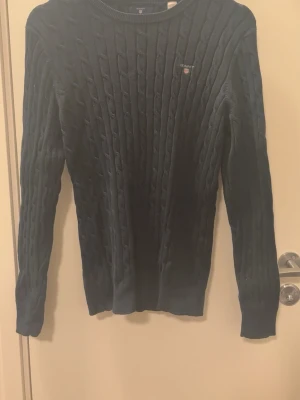 Mörkblå kabelstickad tröja GANT - Mörkblå kabelstickad tröja från GANT med rund halsringning och diskret logga broderad på bröstet. Tröjan har långa ärmar och ribbade muddar vid ärmslut och nederkant. Perfekt för en stilren och klassisk look.