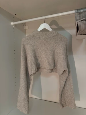 Beige croppad stickad tröja Monki - Trendig beige croppad stickad tröja från Monki i storlek XS. Tröjan har breda ärmar och ribbad halsringning, perfekt för att skapa en cool lager-på-lager-look. Mjuk och skön kvalitet som passar till många outfits.