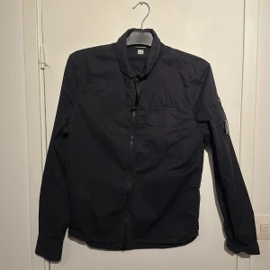 Mörkblå overshirt från C.P. Company - Mörkblå overshirt från C.P. Company med ikonisk linsdetalj på ärmen. Jackan har dragkedja framtill. Den är välanvänd men inga defekter.