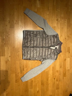 Äkta Grå Moncler cardigan - Säljer en grå Moncler jacka med quiltad front i mörkgrått och ärmar i mjuk bomull. Öppen för prisförslag vid snabb affär. Jackan har ribbad krage med röd och vit detalj, dragkedja framtill och Moncler-logga på ärmen. Skicka gärna prisförslag! Finns några små defekter men absolut ingen som påverkar. Är storlek L men passar mindre, är 180 och den passar bra. Självklart äkta och finns äkthetsbevis!