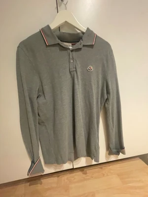 Grå långärmad piké från Moncler - Snygg grå långärmad pikétröja från Moncler med klassisk krage och knappar framtill. Tröjan har Moncler-logga på bröstet och detaljer i rött, vitt och blått på krage och ärmslut. Perfekt för en clean och stilren look.