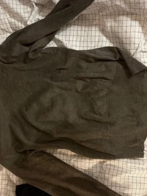 Brungrå  polotröja i mjukt material - Säljer en brungrå polotröja med lång ärm och ribbade muddar. Tröjan har en klassisk, tight passform och är tillverkad i ett tunt, mjukt material som känns skönt mot huden. Perfekt att styla med jeans eller kjol för en clean look.