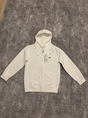 Vit zip hoodie från Lacoste - Snygg vit hoodie från Lacoste med dragkedja framtill och klassisk grön krokodil-logga på bröstet. Hoodien har justerbar huva med snören och två fickor framtill. Tillverkad i mjukt material som känns skönt mot huden. Perfekt för en clean och sportig look.