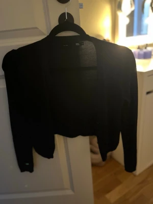 Svart kort kofta från lager 157 - Snygg svart kort kofta från lager 157 med lång ärm och öppen front. Perfekt att slänga över en topp eller klänning för en stilren look. Tunn och lätt i materialet, vilket gör den enkel att bära året runt.