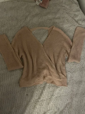 Stickad rosa tröja - Säljer en beige stickad tröja med omlott-design och djup v-ringning. Tröjan har långa ärmar och är tillverkad i ett mjukt, luftigt material som passar perfekt för lager på lager. Enkel och stilren modell som är lätt att matcha, har även öppen rygg bara att skriva om man vill se mer av den