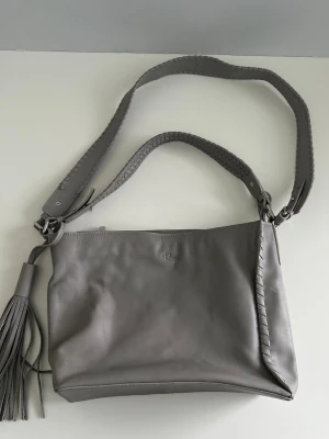 Grå skinnväska från Rizzo - Helt NY grå väska / handväska / axelremsväska i skinn från Rizzo. Praktisk justerbar axelrem och dekorativa inflätade detaljer längs sidorna och handtaget. Dekorativ klassisk borttagbar tofs på sidan. Rymligt inre med flera praktiska fack. Nypris 1500:-, säljes pga felköp.   Hämtas i Stockholm eller skickas mot porto.