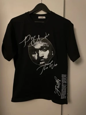 Svart VLONE Pop Smoke t-shirt M - Svart t-shirt från VLONE med Pop Smoke-tryck framtill och stor vit V på ryggen. Snygga vita texter och detaljer på framsidan. Klassisk passform och rund hals. Perfekt för dig som gillar streetwear och vill sticka ut.
