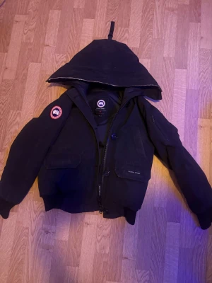 Svart bomberjacka från Canada Goose - Säljer en svart bomberjacka från Canada Goose med huva och ikonisk patch på ärmen. Jackan har dragkedja, knappar och flera fickor framtill. Perfekt för kyligare dagar och har en klassisk, sportig look.