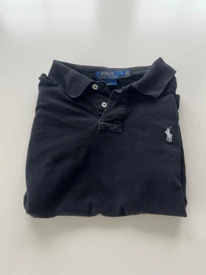 Svart pikétröja från Ralph Lauren - Klassisk svart pikétröja från Ralph Lauren med vit broderad logga på bröstet. Tröjan har krage, knappar framtill och korta ärmar. Tillverkad i mjuk bomull som känns skön mot huden. Perfekt till jeans eller shorts för en clean look.