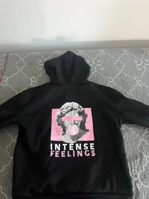 Svart hoodie med konsttryck och text - Snygg svart hoodie med huva och dragsko. På framsidan finns ett stort konstnärligt tryck i rosa och vitt med en staty och texten 'INTENSE FEELINGS'. Hoodien har en klassisk passform och är tillverkad i mjukt material, perfekt för en avslappnad streetwear-look.