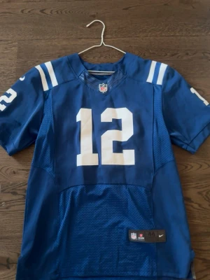 Blå NFL tröja Nike nr 12 - Snygg blå NFL-tröja från Nike med nummer 12 i vitt på bröstet och ärmarna. Tröjan har korta ärmar, vita ränder på axlarna och meshpaneler nedtill för extra ventilation. Perfekt för dig som gillar amerikansk fotboll och vill sticka ut på matchen.