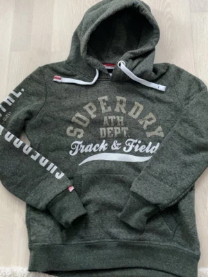 Grön Superdry hoodie med tryck - Mörkgrön hoodie från Superdry med stort tryck på bröstet och ärmen. Klassisk huva med vita dragsnören och känguruficka framtill. Mjuk bomullsblandning och ribbade muddar vid ärmslut och nederkant. Perfekt för en sportig och avslappnad stil.