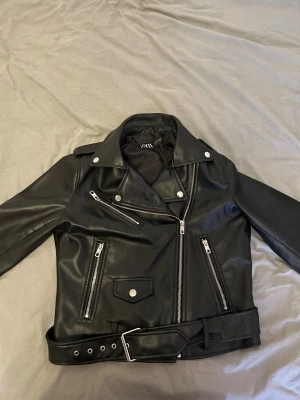 Svart biker skinnjacka från Zara - Snygg svart biker skinnjacka från Zara med silvriga dragkedjor, knappar och bälte i midjan. Klassisk modell med flera fickor och coola detaljer. Perfekt för dig som vill ha en edgy look.