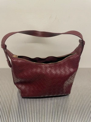 Vinröd flätad handväska i skinn - Snygg vinröd handväska med flätad design och bred axelrem. Väskan har dragkedja upptill och är tillverkad i skinn. Perfekt för dig som vill ha en stilren och trendig accessoar.