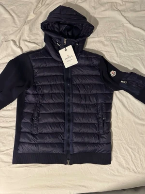 Mörkblå dunjacka med huva från Moncler - Snygg mörkblå dunjacka från Moncler med quiltad front, stickade ärmar och huva. Jackan har dragkedja framtill, två fickor med dragkedja och Moncler-logga på ärmen. Perfekt för kyliga dagar och riktigt stilren look.NFC FUNKAR PRIS KAN DISKUTERAS 