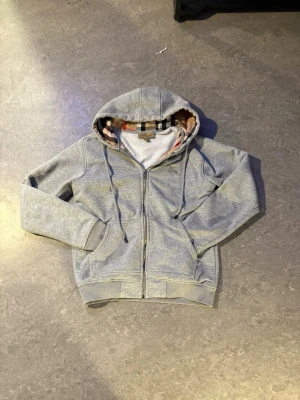 Burrberry hoodie - Snygg grå hoodie med dragkedja från Burberry. Insidan av huvan och öppningen har det klassiska rutmönstret i beige, svart och rött. Hoodien har fickor fram, ribbade muddar och snören i huvan. Perfekt för en chill och stilren look.