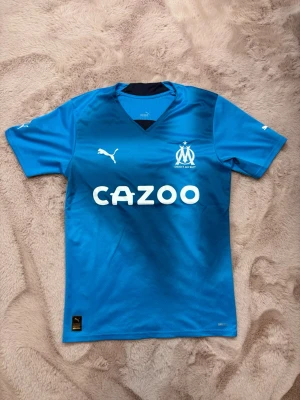 Olympique Marseille Puma - Snygg blå matchtröja från Olympique Marseille med Puma-logga och Cazoo-tryck på bröstet. Tillverkad i lätt och ventilerande funktionsmaterial, perfekt för fotboll eller träning. Klassisk passform med korta ärmar och klubbmärke på bröstet.