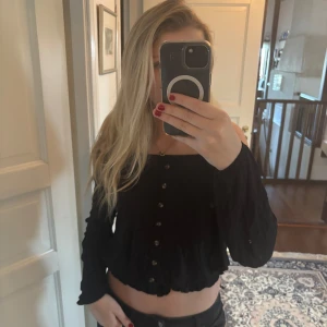 Svart croppad blus med knappar - Trendig svart croppad blus med långa ärmar och knappar framtill. Blusen har en offshoulder-design och en lätt vågig nederkant. Perfekt för dig som gillar en stilren och modern look.