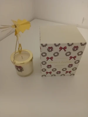 Doftljus med snurrande karusell - Säljer ett doftljus i glasburk med guldfärgat lock och snurrande karusell i metall. Ljusets förpackning är dekorerad med julkransar och röda rosetter. Perfekt som mysig dekoration under vintern eller till jul. Nypris 499:-