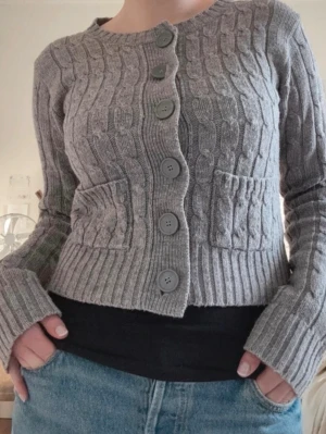 Grå kabelstickad kofta Gina Tricot - Grå kabelstickad kofta från Gina Tricot med stora knappar framtill och två fickor. Koftan har rund halsringning, ribbade muddar och är gjord i stickat material. Jätte gullig o fint skick! 🤗